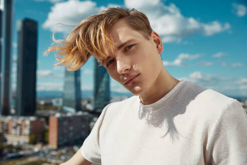 Handsome young male posing while leaning on railing and looking at camera slanted head on blue sky and tall buildings background on sunny day - ADSF38858