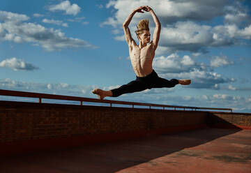 Masterful jungen blonden Balletttänzerin in schwarzen Trikot Durchführung spannende Sprung mit Anhebung Hände auf dem Dach des Gebäudes auf Himmel Hintergrund in sonnigen Tag - ADSF38869