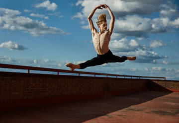 Masterful young blond ballet dancer in black leotard performing exciting jump with raising hands up on roof of building on sky background in sunny day - ADSF38869