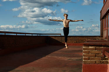 Masterful jungen blonden Balletttänzerin in schwarzen Trikot Durchführung spannende Sprung mit Anhebung Hände auf dem Dach des Gebäudes auf Himmel Hintergrund in sonnigen Tag - ADSF38870