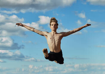 Meisterhafte junge blonde Balletttänzerin in schwarzem Trikot, die einen aufregenden Spagatsprung ausführt und dabei die Hände in die Höhe reckt, vor einem sonnigen Tag - ADSF38872
