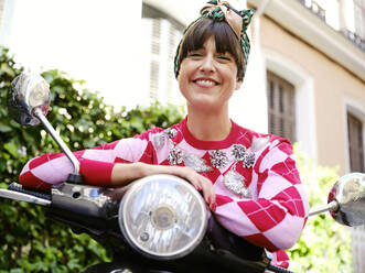 Low angle of smiling woman in vintage outfit sitting on retro motorbike and cheerfully looking at camera - ADSF38876