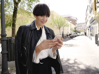 Smiling female with bob hairstyle and in trendy jacket and blouse standing on sidewalk on sunny day and messaging online via cellphone - ADSF38879