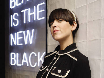 Female model in black wear leaning on wall with illuminate signboard with inscription black is the new black and looking at camera - ADSF38883