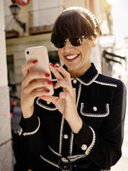 Smiling female with bob hairstyle and in trendy jacket and blouse standing on sidewalk on sunny day and messaging online via cellphone - ADSF38884