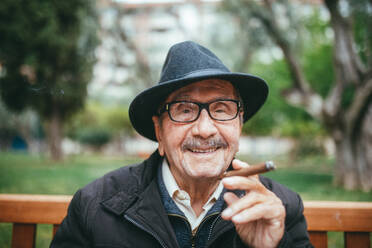Fröhlicher älterer Mann mit Schnurrbart, der einen Hut trägt, Zigarre raucht und in die Kamera schaut, während er auf einer Bank im Park mit grünen Bäumen im unscharfen Hintergrund sitzt - ADSF38896
