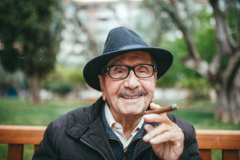 Cheerful senior male with mustache wearing hat smoking cigar and looking at camera while sitting on bench in park with green trees on blurred background - ADSF38896