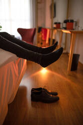 Crop tired male lying on comfortable white bed with shoes off in hotel room with table and armchair near window - ADSF38900