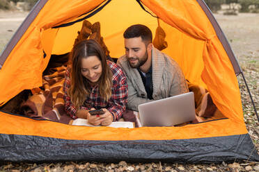 Young couple camping in colorful tent browsing on laptop and smartphone - ADSF38908