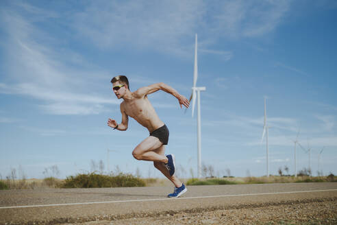 Shirtless athlete running on road by wind turbines - MTBF01258