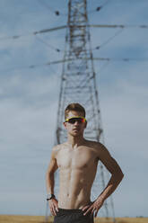 Shirtless young man with hands on hip in front of electric pylon - MTBF01262
