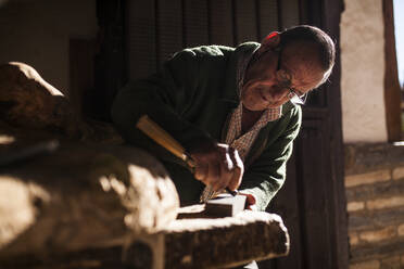 Älterer Holzarbeiter mit Brille bei der Arbeit mit einem Stück Holz in der Werkstatt - ADSF38912