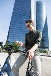 Trendy male standing with modern bike in street on sunny day in downtown while using her smartphone - ADSF38919