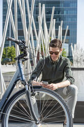 Happy trendy male standing with modern bike in street on sunny day in downtown while using her smartphone - ADSF38920