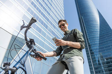 Von unten trendige männliche stehend mit modernen Fahrrad in der Straße auf sonnigen Tag in der Innenstadt, während mit ihrem Smartphone - ADSF38924