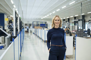 Lächelnde blonde Geschäftsfrau mit Händen in den Taschen in einer Fabrik - JOSEF13295