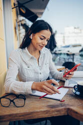 Lächelnde Geschäftsfrau mit Notizblock und Smartphone in einem Straßencafé - OYF00784