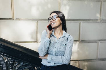 Happy businesswoman talking on smart phone standing in front of wall - OYF00786