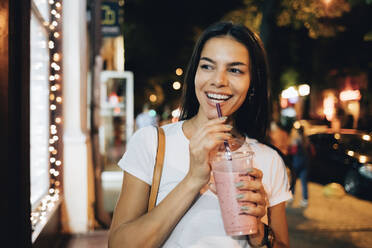 Cheerful woman with smoothie walking on footpath at night - OYF00792