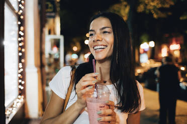 Cheerful woman with smoothie standing at footpath - OYF00795