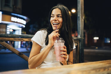 Cheerful woman with smoothie sitting in sidewalk cafe at night - OYF00800