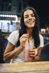 Happy woman holding smoothie sitting at table in sidewalk cafe - OYF00802