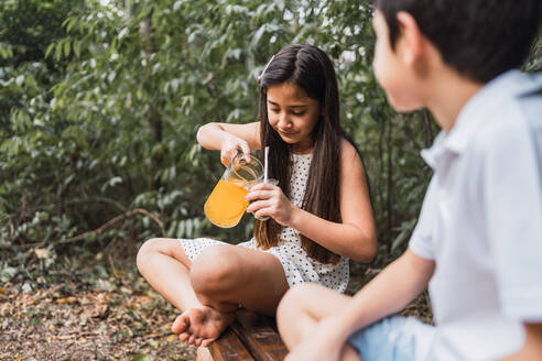 Fröhliches Kind gießt Limonade in eine Kalebasse, während es mit gekreuzten Beinen gegen ein zufriedenes Geschwisterchen auf einer Bank sitzt - ADSF38953