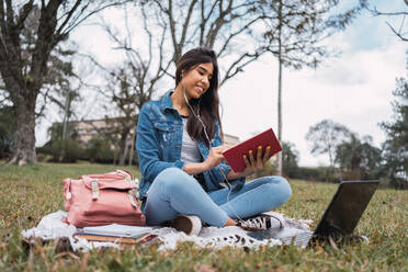 Positive junge hispanische Studentin in Jeanskleidung mit Laptop und Kopfhörern, die Musik hört und ein Buch liest, während sie auf einer grünen Wiese im Campus-Park Hausaufgaben macht - ADSF38957