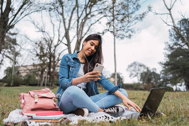 Positive junge hispanische Studentin im Jeans-Outfit, die mit Laptop und Büchern im Park auf ihrem Smartphone surft und online lernt - ADSF38958