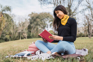 Positive junge hispanische Studentin in Jeanskleidung, die auf einer grünen Wiese im Campus-Park ein Buch liest und Hausaufgaben macht - ADSF38961