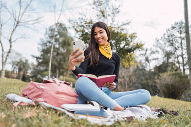 Fröhliche junge hispanische Studentin, die einen Videoanruf auf einem Mobiltelefon tätigt, während sie mit einem offenen Buch auf einer grünen Wiese im Park lernt - ADSF38962