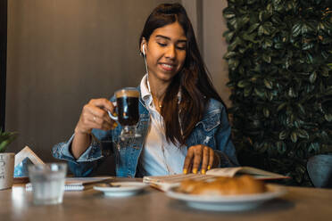 Glückliche junge hispanische Studentin, die Kaffee trinkt und mit Kopfhörern Musik hört, während sie in einem Café ein Buch zur Prüfungsvorbereitung liest - ADSF38963