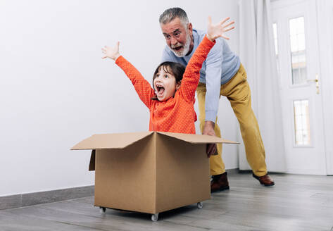 Happy kid and casual clothes pretending to be racer while sitting in carton box while grandfather in glasses pushing box in light room with white walls near door - ADSF38968