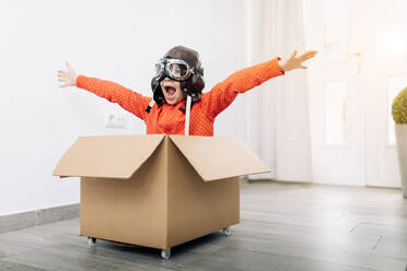 Little kid wearing goggles and aviator helmet while sitting in carton box and pretending to be pilot and looking away an open arms in light room near white door and potted plant - ADSF38970