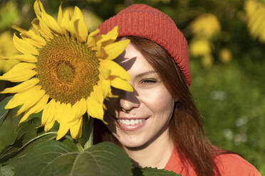 Lächelnde Frau mit Strickmütze hält Sonnenblume an einem sonnigen Tag - VBUF00189