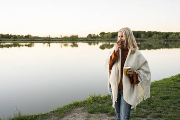 Woman holding cup eating apple by lake at sunset - SVKF00558