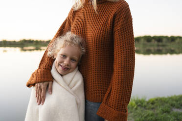 Lächelndes, in eine Decke eingewickeltes Mädchen steht mit seiner Mutter am See - SVKF00560