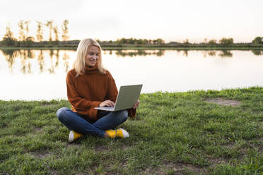 Blonde Freiberuflerin arbeitet am Laptop im Schneidersitz im Gras am See - SVKF00561