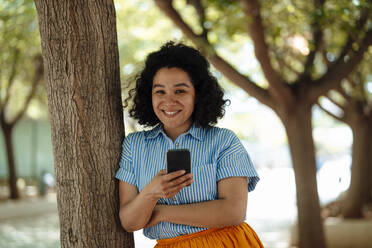 Happy woman with mobile phone leaning on tree trunk - JOSEF13325