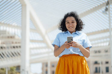 Smiling woman with mobile phone standing on sunny day - JOSEF13330