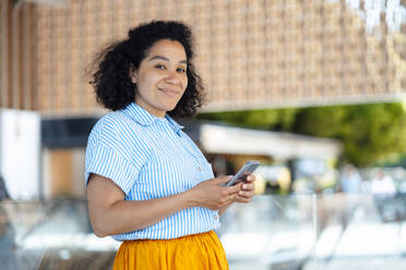 Smiling woman with smart phone leaning on glass railing - JOSEF13333