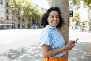 Smiling woman with mobile phone leaning on tree trunk - JOSEF13336