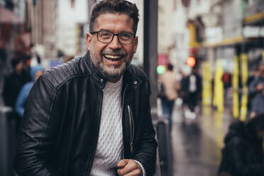 Smiling handsome middle aged man in glasses and leather jacket looking at camera while standing in urban street - ADSF38981