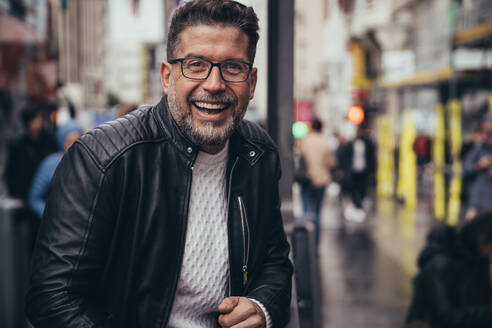 Smiling handsome middle aged man in glasses and leather jacket looking at camera while standing in urban street - ADSF38981