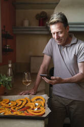 Smiling mature man looking at mobile phone by tray with pumpkin slices and wineglass in kitchen - SVKF00583