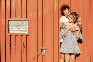 Fröhliches junges gemischtrassiges Paar, das sich zärtlich umarmt, während es auf dem Bürgersteig vor der rot gestreiften Wand eines alten Gebäudes in der Stadt steht - ADSF38986