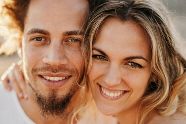 Blonde joyful woman hugging trendy curly man from the back with adoration while looking at camera standing against blurred hills at horizon in sunny evening - ADSF38989