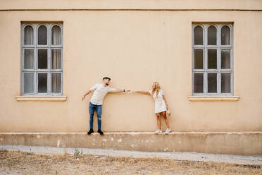 Cheerful adult man and woman in casual clothes with arm raised standing against exterior of building with shabby beige wall and gray windows and holding hands in old town - ADSF38991