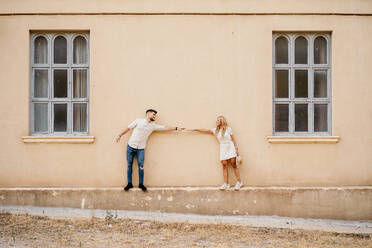 Fröhlicher erwachsener Mann und Frau in Freizeitkleidung mit erhobenem Arm, die an der Außenseite eines Gebäudes mit schäbiger beiger Wand und grauen Fenstern stehen und sich an den Händen halten, in einer alten Stadt - ADSF38991