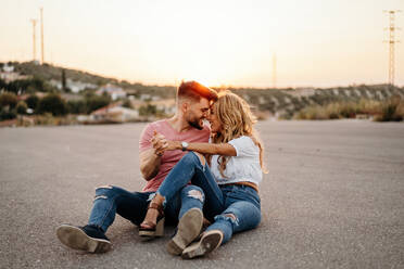 Confident bearded man and magnificent sensual woman in trendy casual clothes embracing while sitting on asphalt road and looking at each other against blurred countryside at dusk - ADSF38997